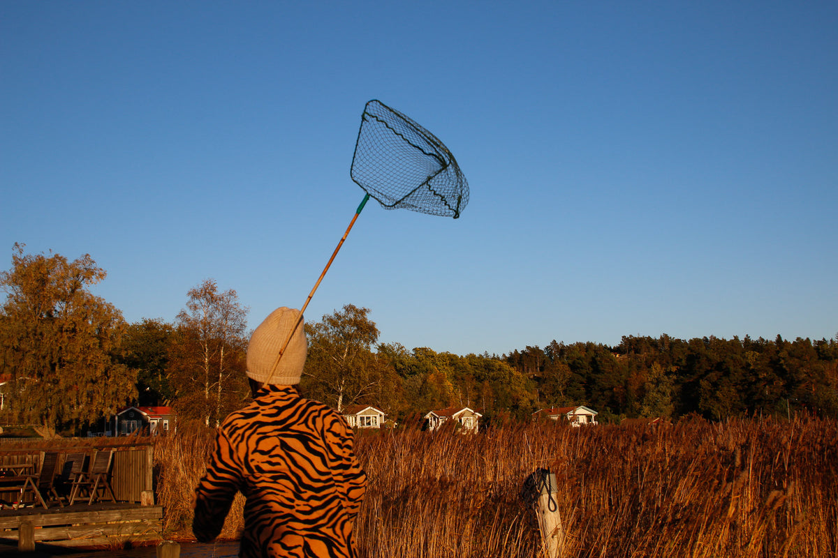En person i en skjorta med tigermönster och en beige mössa håller ett stort håvnät över ett sankt fält i solnedgången, med träd, hus och en klarblå himmel i bakgrunden.